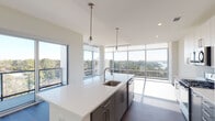 This contemporary 3-bedroom, 3-bathroom residence in Spring, TX offers a well-appointed single-floor layout with striking modern design elements throughout. The open-concept living and kitchen area is a standout, featuring floor-to-ceiling glass windows and sliding doors that flood the space with natural light and frame sweeping panoramic views of the surrounding treetops, waterway, and skyline. The kitchen is equipped with stainless steel appliances, a gas range, a French-door refrigerator, and a large center island with a white quartz countertop, undermount sink, and stylish pendant lighting. White upper cabinetry pairs with gray lower cabinets for a clean, modern aesthetic. The bedrooms are carpeted and generously sized, each offering walk-in closets with built-in shelving. The bathrooms feature dark tile flooring and contemporary vanities, maintaining a consistent modern palette throughout. An in-unit laundry closet houses a full-size washer and dryer. Dark hardwood-style flooring flows through the main living areas, complementing the light-toned walls and recessed lighting. The overall ambiance is bright, clean, and contemporary, with abundant natural light and expansive outdoor views defining the character of this elevated residence.