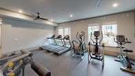 This comprehensive fitness center is thoughtfully designed across multiple interconnected zones, each tailored to a specific aspect of training. The cardio area features a row of treadmills and elliptical machines positioned alongside large windows that allow ample natural light and outdoor views, creating an energizing environment for endurance workouts. Ceiling fans overhead enhance airflow and comfort throughout the space.

The strength training zone is outfitted with a robust selection of free weights, weight benches, cable machines, and plate-loaded equipment, all set on durable rubber flooring ideal for heavy use. Motivational murals and nature-inspired wall graphics add visual energy to this area, complemented by wall-mounted televisions.

A dedicated functional training section houses specialty equipment including a Jacob's Ladder climber, battle ropes, TRX suspension trainers, and a heavy punching bag, catering to high-intensity and cross-training workouts.

Adjacent to the main workout areas is a flexible open studio space featuring patterned carpet flooring, a ceiling fan, and built-in dark shelving units stocked with yoga mats, balance equipment, and accessories. Glass-panel doors connect this area to the rest of the facility. Recessed lighting throughout ensures consistent illumination across all zones, contributing to a polished, modern fitness environment.
