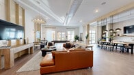 Located at 1647 E Colonial Dr in Orlando, Florida, this single-floor amenity space offers a generously sized, open-concept layout designed for both socializing and relaxation. The interior is defined by clean white walls, warm wood-look flooring, and a striking linear ceiling detail that draws the eye across the expansive living area. Large windows and glass French doors flood the space with abundant natural light while providing direct views of and access to the surrounding outdoor courtyard. The living room area is anchored by a comfortable seating arrangement and a wall-mounted flat-screen TV, complemented by a billiards table that adds a recreational focal point. A sculptural pendant light and bold wall art contribute to the space's contemporary aesthetic. The kitchen features a streamlined white countertop with a brass-finish faucet, textured tile backsplash set against an exposed concrete block wall, open wood shelving, and stainless steel appliances including a dishwasher and undercounter refrigerator. A long communal dining table with modern pendant lighting sits adjacent to the kitchen, creating a cohesive area for shared meals and collaboration. The space is accessed through a sleek steel-and-glass partition door, reinforcing the modern, design-forward character of this well-appointed communal retreat.