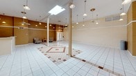 This single-floor space offers a generously proportioned open hall designed to accommodate a variety of uses, from community gatherings to events. The main area is anchored by gleaming white ceramic tile flooring adorned with a distinctive inlaid geometric border pattern in terracotta brown, adding a decorative focal point to the expansive floor plan. Structural columns punctuate the open layout while preserving the sense of spaciousness throughout.

The soaring vaulted ceilings are fitted with a large skylight that floods the interior with natural light, complemented by an array of pendant lights suspended at varying heights to provide warm, even illumination throughout the space. The walls feature painted exposed brick in a warm tan hue, lending a classic, durable character to the interior, with a painted white horizontal trim band adding architectural definition.

A dedicated kitchen area is accessible from the main hall and is equipped with white cabinetry, a full suite of appliances including a refrigerator, stove, microwave, and dishwasher, along with a countertop pass-through that connects seamlessly to the main space. A separate utility alcove offers additional functional space. The front entry door provides a welcoming access point, and the overall layout lends itself well to flexible configuration for a wide range of uses.