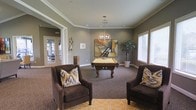 This single-floor amenity space combines recreational and lounge functions in a thoughtfully designed interior. The room centers around an ornately carved wooden billiard table set beneath a globe chandelier that serves as a striking focal point. Crown molding frames the ceiling, adding a refined architectural detail that complements the neutral gray walls and plush, patterned carpet underfoot. A vibrant yellow geometric area rug anchors the seating arrangement, where two deep brown upholstered armchairs with bold ikat throw pillows invite comfortable conversation. Natural light filters in through a large window dressed with white blinds, brightening the space alongside recessed ceiling lighting. Abstract wall art and decorative metallic wall accents contribute to the contemporary, curated aesthetic. A tall potted plant in the corner adds a natural element. Notably, a glass-paned door provides direct access to an adjacent fitness room, visible in the background, making this space part of a larger amenity suite. The overall ambiance is sophisticated yet approachable, well-suited for social gatherings and leisure activities.
