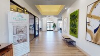 This single-floor residential clubhouse and leasing center presents a polished, contemporary design throughout its well-appointed spaces. The grand entrance corridor sets an immediate tone, featuring light wood-look flooring, a coffered ceiling with a warm yellow inset panel, and a striking oversized living moss wall that adds a vibrant, natural element against the crisp white walls. Arched architectural details and a decorative chandelier lend the hallway an elegant, inviting character, while large glass-panel doors allow ample natural light to filter through.

Off the main corridor, a leasing and meeting area is enclosed by sleek, dark-framed glass partitions, offering a professional yet open environment. A comfortable lounge area features teal banquette seating, round glass-top tables, and contemporary abstract artwork. A separate sitting room showcases a dramatic marble-surround fireplace with a mounted display, flanked by ornate arched niches with decorative tile insets — a refined architectural focal point. Large windows dressed with sheer drapes bring in natural light and outdoor views.

The game room provides a lively social hub with a full-size pool table, soaring ceilings, a circular statement chandelier, lush potted plants, and a dining table for group gatherings. Throughout, the cohesive blue and teal color palette, curated artwork, and light hardwood flooring create a bright, modern ambiance.