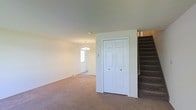 This two-story home presents a welcoming main level characterized by neutral-toned carpeting that extends throughout the open living area. A classic six-panel double closet door provides convenient storage near the entryway, while a carpeted staircase with dark treads leads to the upper floor. The living space is brightened by a modest chandelier that adds a touch of elegance to the room. Natural light filters in through a window near the front entry, contributing to the bright, airy atmosphere of the main level. The walls are finished in a soft, neutral tone, creating a clean and versatile backdrop suited to a variety of interior styles. The layout appears open and functional, with a clear sightline from the living area toward the front door. The overall design is straightforward and practical, offering a comfortable foundation for personalization. The transition between floors is easily accessible via the staircase, suggesting a well-organized vertical layout throughout the home.
