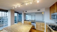 This contemporary single-floor residence showcases a thoughtfully designed open-concept layout that seamlessly blends functionality with modern aesthetics. The kitchen serves as a central highlight, featuring striking granite countertops with rich, flowing veining that anchor the space with a sense of natural elegance. Light wood cabinetry provides a warm contrast to the stone surfaces, while stainless steel appliances, including a built-in dishwasher, add a sleek, professional touch. Track lighting runs along the ceiling, offering adjustable illumination that enhances both task and ambient lighting throughout the kitchen and adjoining living areas. A mirrored backsplash behind the sink cleverly amplifies the sense of depth and light within the space. Hardwood flooring extends across the open floor plan, contributing to the warm and cohesive atmosphere. Perhaps the most notable feature is the abundance of natural light, courtesy of expansive floor-to-ceiling windows and a glass-panel balcony door that frame impressive urban skyline views. The open sightlines and generous glazing create a bright, airy environment that connects the interior living space to the dynamic cityscape beyond.
