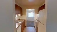 This single-floor space features a galley-style kitchen that serves as the functional heart of the unit. The kitchen is lined on both sides with warm wood-toned upper and lower cabinetry, providing ample storage. A laminate countertop runs along one side, incorporating a stainless steel sink, while white appliances — including a freestanding electric range and a full-size refrigerator — line the opposite wall. The tile flooring in the kitchen offers easy maintenance and a clean, neutral aesthetic.

The kitchen opens directly into an adjoining room with plush carpeting, creating a natural transition between the two spaces. This secondary room benefits from a window that brings in natural light, and a ceiling fan with a light fixture is centrally mounted overhead, providing both ventilation and ambient lighting. The warm beige wall tones throughout create a cohesive, understated atmosphere. The straightforward layout and practical finishes make this space functional and easy to navigate, with a clear sense of flow between the kitchen and the adjoining living area.