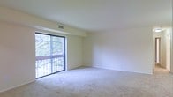 This single-floor space features an open living area finished with wall-to-wall carpeting in a neutral tone, contributing to a clean and understated aesthetic. The room is characterized by smooth, light-colored walls and a white ceiling with a centrally positioned HVAC vent, indicating a built-in climate control system throughout. A large sliding glass door dominates one wall, functioning as both a source of natural light and a direct access point to a private balcony. The balcony overlooks a leafy outdoor setting, with mature trees visible through the glass, providing a natural backdrop and a sense of seclusion. The sliding door is framed by a dark aluminum track and is flanked by a fixed glass panel, maximizing the view and the inflow of daylight. The open layout of the living area offers flexibility for various furniture arrangements. The overall ambiance is calm and neutral, with the combination of soft carpeting, pale walls, and abundant natural light creating a bright and airy environment well-suited for comfortable everyday living.