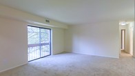 This single-floor space features an open living area finished with wall-to-wall carpeting in a neutral tone, contributing to a clean and understated aesthetic. The room is characterized by smooth, light-colored walls and a white ceiling with a centrally positioned HVAC vent, indicating a built-in climate control system throughout. A large sliding glass door dominates one wall, functioning as both a source of natural light and a direct access point to a private balcony. The balcony overlooks a leafy outdoor setting, with mature trees visible through the glass, providing a natural backdrop and a sense of seclusion. The sliding door is framed by a dark aluminum track and is flanked by a fixed glass panel, maximizing the view and the inflow of daylight. The open layout of the living area offers flexibility for various furniture arrangements. The overall ambiance is calm and neutral, with the combination of soft carpeting, pale walls, and abundant natural light creating a bright and airy environment well-suited for comfortable everyday living.