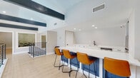 Located at 4615 S Virginia St in Amarillo, TX, this two-story multi-use facility combines modern design with functional amenity spaces. The upper level features a bright, open lounge and kitchen area finished with light hardwood flooring, soaring ceilings accented by bold dark exposed beams, and recessed lighting. A sleek white kitchenette with quartz countertops, stainless steel appliances, a black matte faucet, and ample cabinetry serves the space efficiently. A large island with tan leather barstools provides casual seating, while a separate dining nook with a dark table and vibrant red chairs adds a lively touch. A cozy lounge corner anchors the upper level with a tile-surround fireplace, a shiplap accent wall, original artwork, exposed beam ceilings, and comfortable seating arranged around a copper-topped coffee table. A modern open-railing staircase connects the two floors. The lower level houses a well-appointed fitness center with treadmills, stationary bikes, weight equipment, and a dedicated open stretching room — all set against energetic teal and yellow striped walls with carpet flooring. A vending machine adds everyday convenience. The overall ambiance blends contemporary finishes with a welcoming, active-community atmosphere.