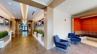 This single-floor amenity space presents a thoughtfully designed environment that blends modern aesthetics with practical functionality. The main corridor serves as the spine of the space, finished with large-format marble-look tile flooring and warm wood-paneled columns that frame the journey through the building. Decorative globe pendant lighting adds an elegant touch overhead, while built-in planters with ornamental grasses introduce natural elements along the hallway.

The welcoming lobby and leasing area features floor-to-ceiling glass partitions with sleek black steel framing, allowing visual connectivity throughout. A bold orange sculptural "RENT" sign and a wall-mounted display screen provide branded focal points, while a coffered ceiling with a statement pendant light anchors the entry experience.

A glass-enclosed lounge or club room, accessible via wood-framed sliding glass doors flanked by built-in shelving, offers a vibrant social setting with colorful furnishings and ample seating. Adjacent spaces include a fully equipped catering kitchen outfitted with stainless steel appliances—including a French door refrigerator, double wall oven, and dishwasher—paired with light wood cabinetry and stone-look tile flooring.

The fitness center, visible in the dollhouse view, is well-appointed with a comprehensive array of exercise equipment. Throughout, the design maintains a consistent palette of warm wood tones, crisp white walls, and polished tile, creating a cohesive, upscale atmosphere.