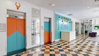 Located at 111 Cooper St in Ottawa's Centretown neighborhood, One Eleven is a dynamic, single-floor amenity space that makes a bold design statement from the moment you step inside. The entrance greets visitors with a striking multi-toned checkered marble floor, eye-catching teal accent walls, and color-blocked elevator doors in vivid orange and teal — all anchored by the prominently displayed "One Eleven" signage at a welcoming reception desk. The space extends generously, offering a seamless flow between distinct zones. The kitchen and living area features high-top communal tables with vibrant orange and red stools, stylish pendant lighting, built-in shelving, and wall-mounted televisions — ideal for both casual dining and social gatherings. Decorative patterned floor tiles transition throughout, adding visual character and energy. The game room is a standout highlight, equipped with multiple ping-pong tables, teal and purple lounge seating, bold geometric wall panels, and vending machines, all bathed in natural light from large windows overlooking greenery. Cozy reading nooks with plush sofas and built-in bookshelves offer quieter retreats. The overall ambiance is lively, youthful, and thoughtfully designed, combining functionality with an unmistakably vibrant aesthetic throughout.