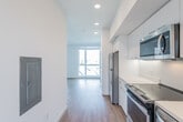 This contemporary single-floor unit offers a thoughtfully designed layout with a clean, modern aesthetic carried throughout every room. The galley-style kitchen is a standout feature, outfitted with a full suite of stainless steel appliances including a refrigerator, electric range, built-in microwave, and dishwasher. Crisp white cabinetry extends from counter height to the ceiling, maximizing storage, while white quartz-style countertops and a stainless steel sink complete the polished look. Warm hardwood-style flooring runs continuously throughout the space, adding a sense of cohesion and warmth against the bright white walls and recessed lighting above. The open living area is generously proportioned, with large windows that invite abundant natural light and offer views of the surrounding neighborhood. A wall-mounted mini-split unit provides efficient climate control. A louvered interior door adds a subtle design detail between spaces. The bathroom is clean and functional, featuring a full soaking tub and shower combo with white tile surround, chrome fixtures, and recessed lighting. Overall, the unit presents a bright, well-appointed, and move-in-ready space with a cohesive modern design language throughout.