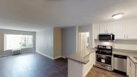 This single-floor residence offers a clean, modern aesthetic throughout its open and functional layout. The kitchen is a standout feature, outfitted with crisp white shaker-style cabinetry, stainless steel appliances including a gas range and over-the-range microwave, and a peninsula countertop with a neutral gray surface that serves as both a prep area and casual dining space. The kitchen flows seamlessly into the living area, creating an open-concept environment ideal for entertaining or everyday living. Rich, dark hardwood-style flooring runs throughout the main living spaces, providing a warm contrast to the cool-toned gray and beige walls. Natural light filters in through a window in the living area, enhancing the bright and airy feel of the space. The neutral color palette, clean lines, and contemporary finishes give the interior a fresh, move-in-ready appeal. A doorway visible from the main living area leads to an additional room, hinting at a practical and well-organized floor plan. The smooth white ceilings and crisp white trim complete the polished, cohesive look of this thoughtfully designed residence.