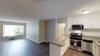 This single-floor residence offers a clean, modern aesthetic throughout its open and functional layout. The kitchen is a standout feature, outfitted with crisp white shaker-style cabinetry, stainless steel appliances including a gas range and over-the-range microwave, and a peninsula countertop with a neutral gray surface that serves as both a prep area and casual dining space. The kitchen flows seamlessly into the living area, creating an open-concept environment ideal for entertaining or everyday living. Rich, dark hardwood-style flooring runs throughout the main living spaces, providing a warm contrast to the cool-toned gray and beige walls. Natural light filters in through a window in the living area, enhancing the bright and airy feel of the space. The neutral color palette, clean lines, and contemporary finishes give the interior a fresh, move-in-ready appeal. A doorway visible from the main living area leads to an additional room, hinting at a practical and well-organized floor plan. The smooth white ceilings and crisp white trim complete the polished, cohesive look of this thoughtfully designed residence.