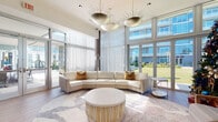 This elegantly designed single-floor amenity lounge offers a refined and welcoming atmosphere, combining contemporary architecture with thoughtfully curated interiors. The space is anchored by soaring ceilings and expansive floor-to-ceiling windows that flood the interior with natural light and frame sweeping outdoor views, including lush greenery and a neighboring skyline. Wide-plank hardwood flooring flows throughout, grounding the bright, airy aesthetic. The main lounge area features a generous curved sectional sofa in warm cream tones, layered over a sculptural area rug, and crowned by striking sunburst-style pendant chandeliers that serve as artistic focal points. Large bi-fold glass doors open directly to an outdoor patio, offering seamless indoor-outdoor connectivity. Sheer floor-to-ceiling drapery softens the wall of glass, adding an element of understated luxury. A separate gathering zone provides additional seating with coordinated accent chairs and cylindrical side tables, ideal for casual socializing. The wet bar area showcases rich wood cabinetry, a iridescent penny-tile backsplash, a granite countertop, and bar stool seating, complemented by an artisan pendant light fixture overhead. A private dining nook with floor-to-ceiling windows offers panoramic city views, while glass-partitioned rooms allow for flexible, semi-private use. Throughout, the design balances warmth, openness, and modern sophistication.