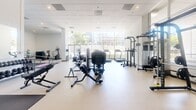 Located on Wilshire Boulevard in the heart of Los Angeles, this fitness center offers a thoughtfully designed, single-floor exercise space that accommodates a wide range of workout needs. The open floor plan is anchored by soaring ceilings and a dramatic wall of floor-to-ceiling windows along one side, flooding the interior with abundant natural light and offering views of the surrounding cityscape.

The exercise room is equipped with a comprehensive selection of cardio machines, including treadmills, ellipticals, and stationary bikes, all positioned to take advantage of the natural light from the expansive window wall. A dedicated strength training area features a variety of cable machines, free weights, and multi-functional racks, providing options for users at all fitness levels. A large padded mat area with yoga mats offers space for stretching, yoga, or floor-based exercises.

The interior design is clean and contemporary, with white walls, light-toned flooring, and a distinctive accent wall featuring a playful, repeating dumbbell graphic in pink and teal — adding a vibrant, energetic character to the space. A rack of medicine balls and additional accessories are neatly organized near the entry, which is accessible via a step and ramp entry with glass doors. The overall ambiance is bright, modern, and motivating.