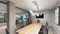This contemporary amenity space seamlessly combines a professional conference room with an adjacent fitness center, offering a well-rounded environment for both work and wellness. The boardroom is anchored by a substantial natural wood conference table surrounded by high-back ergonomic chairs, creating a polished setting for meetings and collaboration. A wall-mounted flat-screen display provides built-in presentation capabilities, while the room's standout feature is a sculptural, free-flowing LED light fixture suspended from the ceiling — an artistic centerpiece that adds a dynamic visual element to the otherwise clean, neutral aesthetic. The ceiling features integrated linear ventilation, contributing to a refined and uncluttered look. Floor-to-ceiling glass partition walls separate the conference room from the fitness center, maintaining an open, transparent feel while visually connecting both spaces. The gym, visible through the glass, is outfitted with cardio equipment and benefits from its own generous lighting. Light-toned walls and warm wood finishes throughout create a balanced, professional ambiance that feels both functional and design-forward.