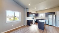 Located at 12501 W Sixth Ave in Airway Heights, Washington, this single-floor residence offers a thoughtfully designed layout with modern finishes and an airy, open feel. The kitchen is a standout feature, showcasing rich dark cabinetry with sleek hardware, crisp white countertops, a central island for additional prep space, and a full suite of stainless steel appliances including a range, microwave, refrigerator, and double-basin sink. Natural light pours in through multiple windows, brightening the warm wood-look luxury vinyl plank flooring that runs throughout the main living and kitchen areas. The living space impresses with dramatically tall ceilings and clerestory windows that amplify the sense of volume, complemented by a sliding glass door that invites natural light and outdoor views. Bedrooms are comfortably sized, finished with plush carpeting, neutral wall tones, and bright windows. The bathroom is cleanly appointed with a full tub-shower combination and chrome fixtures. Throughout the residence, consistent neutral paint tones, white panel doors, and clean architectural lines create a cohesive, contemporary aesthetic suited for comfortable everyday living.
