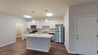This single-floor unit in Nashville, Tennessee offers a clean, contemporary design with a neutral color palette and durable finishes throughout. The open-concept layout connects the living area and kitchen seamlessly, with rich dark hardwood-style flooring anchoring the shared spaces. The kitchen is well-equipped with stainless steel appliances, including a refrigerator, electric range, microwave, and dishwasher. White shaker-style cabinetry extends along the full wall, providing generous storage, while light stone-look countertops add a polished touch. A center island offers additional prep space and casual seating potential. Pendant lighting above the island and recessed fixtures illuminate the area effectively. The living area is open and spacious, providing a flexible layout for various furniture arrangements. The bedrooms feature plush wall-to-wall carpeting, natural light from windows with blinds, and ceiling fans for added comfort. The bathrooms maintain the same cohesive aesthetic, with white vanity cabinetry, stone-look countertops, large mirrors, and tub-shower combinations accented by wood-toned tile surrounds and dark hardwood-style flooring, creating a warm yet modern finish.