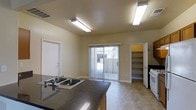 This two-story space offers a functional and open layout, beginning with a combined kitchen and living area on the main floor. The kitchen is equipped with dark granite-style countertops, a double-basin sink, and a white range, providing a practical cooking environment. Fluorescent ceiling fixtures illuminate the space evenly, complementing the neutral beige walls and tile flooring that run throughout the main level.

A sliding glass door floods the area with natural light and provides direct access to an outdoor patio or exterior space, enhancing the overall openness of the room. Adjacent to the kitchen, a dedicated pantry with built-in shelving offers convenient dry storage, keeping the living area uncluttered. The open-concept design between the kitchen and living zones creates a spacious, connected feel suitable for everyday use.

The layout is straightforward and clean, with white-trimmed doorways and minimal architectural detailing that give the space a simple, utilitarian aesthetic. The second floor likely provides additional living or sleeping quarters, expanding the overall functionality of the home across multiple levels.