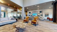 The Reach on Goodale, located at 644 Chlois Lane in Columbus, Ohio, presents a sophisticated and contemporary leasing and amenity lobby designed to make a lasting first impression. The single-floor space is defined by soaring ceilings that amplify the sense of openness, complemented by large floor-to-ceiling windows and glass-panel walls that flood the interior with abundant natural light and offer views of the surrounding landscaping.

Wide-plank hardwood-style flooring runs throughout, unifying the various seating areas that are thoughtfully arranged across the expansive space. Multiple lounge zones feature an eclectic mix of upscale furnishings, including rich cognac leather wingback chairs, plush velvet accent chairs in teal and olive, and neutral sofas arranged around sculptural coffee tables. A bold teal accent wall adds a vibrant focal point near the main entrance, while a decorative media wall with backlit paneling and built-in display niches anchors the primary lounge.

A glass-enclosed leasing office provides a transparent yet defined workspace, maintaining visual connectivity with the lobby. Additional seating alcoves with fireside chairs and curated wall art create intimate gathering spots. The overall design blends warm wood tones, metallic accents, and modern lighting fixtures — including a striking sputnik chandelier — to craft a refined, welcoming atmosphere that reflects the property's upscale character.
