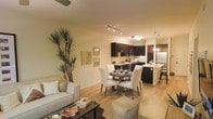 This single-floor residence showcases an open-concept layout that fluidly connects the living, dining, and kitchen areas into one cohesive and functional space. The living area is furnished with a light-toned sofa, a sleek glass-top coffee table, and a tall decorative plant that adds a natural, organic element to the room. Warm beige walls and light hardwood-style flooring run throughout, creating a consistent and inviting atmosphere.

The dining area sits centrally within the space, featuring a round dark wood table surrounded by white upholstered chairs, providing a comfortable and stylish setting for meals. A track lighting fixture overhead illuminates this zone effectively.

The kitchen is defined by dark espresso cabinetry that contrasts sharply with lighter countertops, stainless steel appliances, and an under-cabinet lighting system that adds both function and ambiance. A breakfast bar with pendant lighting extends from the kitchen, offering additional seating and serving as a natural divider between the culinary and dining spaces.

Overall, the space feels cohesive, modern, and well-lit, with a warm and welcoming ambiance suitable for everyday living and entertaining.