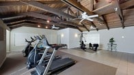 This dedicated fitness space combines rustic architectural character with practical workout functionality. The most striking feature is the dramatic exposed wood beam ceiling, where dark timber rafters and trusses span the full width of the room, adding warmth and a distinctly rustic aesthetic. Multiple ceiling fans are mounted throughout, ensuring comfortable airflow during workouts.

The space is divided into distinct workout zones. The cardio area houses several machines, including treadmills and an elliptical trainer, positioned along one side of the room. Protective rubber flooring underlies the equipment, preserving the surface and providing traction. The strength training zone, situated toward the far end, features a multi-station cable machine and a weight bench, set atop tile flooring that transitions from the rubber matting.

White walls contrast beautifully with the dark wood ceiling, keeping the space feeling open and bright. A large window allows natural light to filter in, complementing the recessed track lighting and wall sconces. A wall-mounted mirror near the cardio equipment enhances the sense of space and supports exercise form. The overall ambiance is both functional and characterful.