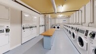 Located at 14412 Seymour St in Halifax, Nova Scotia, this shared laundry facility offers a well-organized and functional space designed to serve multiple users efficiently. The room features a long, rectangular layout with white walls and light grey tile flooring, creating a clean and utilitarian atmosphere throughout.

Along the primary wall, a full row of front-loading washing machines is neatly arranged, providing substantial washing capacity. Opposite and adjacent areas house stacked dryer units, maximizing the use of available wall space. A centrally positioned wooden folding table runs along the middle of the room, offering a practical surface for sorting and folding laundry.

The ceiling is finished in a warm yellow tone and is equipped with exposed ventilation ductwork and piping, along with fluorescent strip lighting that ensures the space is well-lit. Coin-operated and card-based payment kiosks are mounted on the walls, accompanied by posted instructional and informational signage. A waste bin is also conveniently placed within the room.

The layout is thoughtfully arranged to allow users to move comfortably between machines and the folding table. Overall, the space presents a practical, no-frills laundry facility that prioritizes functionality and capacity for shared residential use.