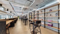 Located at 1 Monroe St in Norwalk, Connecticut, this single-floor commercial space offers a generous open-plan layout with a distinctly industrial-modern aesthetic. The design is defined by exposed concrete ceilings with visible ductwork and piping, polished concrete floors, and raw concrete block walls that lend the space an authentic, loft-style character. Linear LED lighting runs throughout, complementing the natural light that pours in through expansive floor-to-ceiling windows along the perimeter, offering views of the surrounding streetscape. The space is organized into several functional zones. A café-style bar area features open wood shelving mounted on dark steel frames, a service counter with built-in cabinetry, and bar-height seating, creating a casual gathering and refreshment hub. Adjacent breakout areas include high-top tables and curated shelf displays with decorative accessories, ideal for informal meetings or collaborative work. A comfortable lounge zone is furnished with plush seating arrangements—including armchairs and a sofa grouping set on layered rugs—providing a relaxed environment for conversation or focused work. Dark steel partitions and wire-mesh railings define transitions between zones while maintaining visual openness throughout. The overall ambiance is refined yet approachable, balancing industrial raw materials with warm wood accents and thoughtfully curated furnishings.