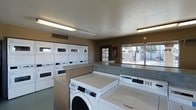 This single-floor shared laundry facility is designed to accommodate the needs of multiple users efficiently and comfortably. The room is furnished with a generous number of commercial-grade washing machines and dryers, including both front-loading washers and stacked dryer units, arranged to maximize capacity and ease of use. Spacious countertops line the walls, providing ample surface area for sorting and folding laundry. The layout is thoughtfully organized, with machines and folding surfaces positioned to allow comfortable movement throughout the space. Large windows along one wall allow natural light to fill the room, creating a bright and welcoming atmosphere while also offering views of the outdoor surroundings. A glass-paneled door provides additional access and visibility to the exterior. Overhead fluorescent lighting ensures the space remains well-lit at all times. The walls are finished in a warm neutral tone, and the smooth ceiling features integrated ventilation to maintain air quality. A fire safety device is mounted on the wall, reflecting attention to safety standards. Overall, this laundry facility is a practical, clean, and functional shared amenity space.