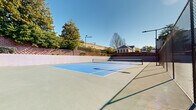 Located at 4835 Cameron Valley Parkway in Charlotte, NC, this outdoor recreational facility offers a well-appointed sports and leisure complex set against a backdrop of mature trees and clear Carolina skies. The centerpiece is a regulation-sized tennis court featuring a vibrant blue and green hard-court surface with crisp white line markings and a centered net, designed to meet the needs of both casual players and serious enthusiasts. The court is enclosed by sturdy chain-link fencing with a gated entry point, ensuring safety and defined play areas. Overhead sports lighting poles are positioned throughout the facility, allowing for evening use after sunset. Adjacent to the tennis court is a generously sized open hardscape area that serves multiple recreational purposes. This space is equipped with an oversized outdoor chess set, providing a unique and engaging activity for residents and visitors of all ages. Benches are thoughtfully placed throughout the area, offering comfortable spots to rest and observe. A nearby building with classic arched architectural detailing and a cupola provides an elegant structural backdrop to the courts. The entire facility is bordered by retaining walls, decorative fencing, and lush greenery, creating an inviting and well-maintained outdoor environment suited for active community recreation.