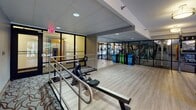 Located at 2500 Clarendon Blvd in Arlington, Virginia, this well-appointed fitness center offers a comprehensive workout environment spread across a single floor. The space is designed with light hardwood flooring throughout, creating a clean and modern aesthetic that ties the various exercise zones together seamlessly.

The functional training area is a standout feature, equipped with a dedicated TRX Training Zone rig stocked with suspension trainers, medicine balls, kettlebells, and resistance equipment neatly organized on multi-tiered metal racks. Large floor-to-ceiling windows flood this section with natural light and offer calming views of the surrounding greenery, creating an energizing atmosphere.

A dedicated cycling studio provides multiple indoor spin bikes arranged for group or individual sessions, complemented by a wall-mounted flat-screen television and warm overhead lighting. This area connects smoothly to an open cardio zone featuring a rowing machine positioned along a railed platform, with reflective glass partitions that visually expand the space.

The strength and cardio equipment room is visible through glass-panel walls, housing a variety of weight machines and additional cardio units. Recessed and flush-mounted lighting throughout ensures consistent illumination across all workout zones. The facility also includes a body weight scale, adding a practical wellness touch. The overall ambiance is professional, clean, and motivating, suited for a wide range of fitness activities.