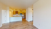 This single-floor residence showcases a thoughtfully designed open-concept layout, connecting the living area seamlessly to a well-appointed kitchen. The kitchen is a standout feature, offering warm-toned flat-front cabinetry, white subway tile backsplash, light countertops, and a full suite of stainless steel appliances including a refrigerator, range, over-the-range microwave, and dishwasher. A peninsula provides additional workspace and serves as a natural divider between the kitchen and living space. Pendant lighting adds a modern touch overhead.

Light wood-style flooring flows consistently throughout the unit, contributing to a cohesive and airy aesthetic. The living area is bright and open, with clean white walls and a neutral palette. A notable highlight is the corner window configuration in one room, featuring floor-to-near-ceiling glass panels that invite generous natural light and offer views of the surrounding neighborhood and tree canopy.

The unit includes two bathrooms, each featuring a tub-shower combination, wood-toned vanity, and large mirror. Storage is well-addressed with a dedicated walk-in closet equipped with a full-width hanging rod and shelf. The overall design is clean, contemporary, and move-in ready.