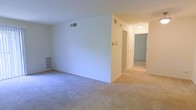 This single-floor residence offers a bright, open layout designed for comfortable everyday living. The main living area is spacious and features soft, neutral wall-to-wall carpeting that extends throughout the space, contributing to a warm and cohesive feel. Crisp white walls reflect natural and artificial light effectively, enhancing the overall sense of openness and cleanliness throughout the interior.

A central hallway connects the living area to additional rooms, providing a practical and well-organized flow through the residence. Overhead ceiling-mounted lighting fixtures illuminate the corridor, ensuring the connecting spaces remain bright and welcoming. HVAC vents are thoughtfully integrated into both the walls and ceilings, reflecting a functional approach to climate control throughout the unit.

Built-in shelving is visible along one wall of the main living area, offering convenient storage or display options without occupying additional floor space. The doorways are clean and simply framed, maintaining the minimalist and understated aesthetic of the interior. The overall ambiance of the space is calm, neutral, and move-in ready, making it a versatile canvas adaptable to a variety of furnishing styles and personal tastes.