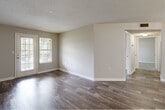 This single-floor unit offers a well-organized layout across several functional spaces, each finished with consistent, neutral tones and clean white trim throughout. The living area is a standout feature, showcasing rich, dark wood-look flooring that extends across much of the unit. Natural light pours in through a set of large windows and a glass-paneled door that opens to an outdoor patio, creating an open and airy atmosphere. The adjacent dining area is highlighted by a classic chandelier and connects seamlessly to the kitchen via a peninsula-style counter with a built-in sink. The galley kitchen is efficiently designed with ample white cabinetry reaching to the ceiling, light-toned countertops, a gas range, and a full-size refrigerator, all set above matching wood-look tile flooring. The bedroom is a comfortable, well-lit retreat featuring plush carpeting and dual windows that overlook lush greenery, providing a peaceful outdoor view. An additional room with wood-look flooring offers flexible use as a second bedroom or office. The bathroom is cleanly appointed with wood-look flooring, a vanity with mirror, and straightforward fixtures. A textured ceiling is consistent throughout the unit, adding a classic residential touch to the overall design.