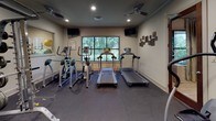 This fitness center offers a comprehensive workout environment designed to support a wide range of exercise routines. The space is finished with durable, dark rubber flooring throughout, providing both safety and functionality for various types of training. Recessed lighting and ceiling fans maintain a comfortable atmosphere, while a large picture window along one wall frames lush outdoor greenery, bringing in natural light and a refreshing view.

The cardio section is well-stocked with multiple treadmills and elliptical machines, each positioned to face the window for an engaging workout experience. Mounted flat-screen televisions are positioned on the walls, offering entertainment during exercise sessions. For strength training, the space includes a multi-station cable machine, a weight rack loaded with plates, and additional resistance equipment to accommodate a variety of lifting routines. A stationary bike and a rowing-style machine round out the equipment selection, ensuring options for both high- and low-impact workouts.

Large wall mirrors expand the sense of space and allow users to monitor their form during exercise. Decorative wall panels add a subtle aesthetic touch. A water cooler is conveniently accessible within the room. Entry is through a glass-paneled wood door, with a pass-through window connecting to an adjacent common area, contributing to the open and accessible feel of this thoughtfully appointed fitness facility.