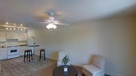 This single-floor space offers a clean, comfortable layout designed for easy living. The living area serves as the heart of the space, featuring neutral-toned walls that create a calm and inviting atmosphere. Two patterned accent chairs are arranged around a dark circular coffee table, providing a simple yet functional seating arrangement. A small potted plant adds a touch of greenery to the area. The room is equipped with a ceiling fan fitted with a multi-bulb light fixture, ensuring both air circulation and adequate lighting throughout the space. The flooring is a warm-toned carpet that complements the overall neutral color palette. Adjacent to the main seating area, a high-top bistro table with bar-height stools offers an additional casual dining or gathering spot. The walls are smooth and finished in a soft off-white, contributing to the bright and airy feel of the room. Baseboards and clean transitions between surfaces reflect a well-maintained interior. Overall, the space presents a straightforward, functional design that prioritizes comfort and ease of use.