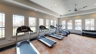 This single-level fitness center offers a comprehensive workout environment thoughtfully arranged to accommodate a variety of exercise needs. The cardio area lines one wall beneath a row of large windows, housing multiple treadmills and elliptical machines that benefit from abundant natural light and outdoor views of the surrounding grounds. Ceiling fans mounted above help maintain airflow throughout the space during use.

The strength training section features a multi-functional cable machine, an adjustable weight bench, and a rack of dumbbells, providing options for resistance-based workouts. A large wall-mounted mirror spans a significant portion of the strength area, visually expanding the room while allowing users to monitor their form. Exercise and medicine balls in various sizes are neatly organized on a dedicated rack nearby.

The flooring features a durable, textured surface in warm terracotta tones, complemented by recessed lighting in the tray ceilings above. The neutral color palette throughout creates a clean, modern aesthetic. With direct exterior access, good ventilation, and a practical layout that separates cardio from strength zones, this fitness center is designed to deliver a functional and motivating workout experience.