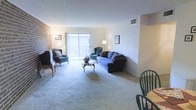 This single-floor residence centers around a spacious, carpeted living room that serves as the heart of the home. The room is furnished with a blue sofa and coordinating wingback chairs arranged around a classic wooden coffee table, creating a comfortable and functional seating area. A prominent exposed brick accent wall adds a touch of architectural character and visual warmth to the otherwise clean, neutral-toned interior.

Natural light fills the living room through a large sliding glass door that opens to an outdoor balcony, offering a pleasant connection to the exterior. The sliding door is framed by light teal curtains, contributing a soft pop of color to the space. Wall-mounted framed artwork and a traditional floor lamp provide tasteful decorative touches.

The ceilings are standard height and include a visible HVAC vent, ensuring climate comfort throughout. The soft, light-toned carpet extends across the living area, adding to the overall sense of warmth and comfort. A dining area is partially visible at the edge of the room, suggesting a practical and well-organized layout. The overall ambiance is classic, understated, and livable.