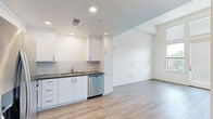 Located at 26701 McBean Pkwy in Santa Clarita, CA, this single-floor unit offers a well-designed layout comprising a living room, kitchen, and bathroom. The living area is a standout feature, characterized by soaring ceilings, crisp white walls, and light wood-tone flooring that extends throughout the space. A full-height glass door flanked by large windows floods the room with natural light and provides direct access to a private balcony with outdoor views, creating an airy, open atmosphere. The kitchen is thoughtfully appointed with white shaker-style cabinetry, dark countertops, a subway tile backsplash, and a full suite of stainless steel appliances including a refrigerator with ice maker, gas range, over-the-range microwave, and dishwasher. Recessed lighting illuminates the kitchen efficiently. The bathroom features a clean, neutral palette with large-format tile surround enclosing a soaking tub and shower combination, complemented by built-in open shelving for convenient storage. Light wood flooring carries into the bathroom as well, tying the overall aesthetic together cohesively. The unit's modern finishes and efficient use of space make it a functional and visually appealing residence in the Santa Clarita Valley.