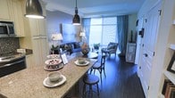 This single-floor space showcases a thoughtfully designed open-concept layout that seamlessly connects the kitchen and living areas. The kitchen features a substantial granite-topped bar counter with seating provided by sleek bar stools, making it an ideal spot for casual dining or entertaining. A bold industrial-style pendant light hangs above the counter, adding a contemporary focal point to the space.

Dark hardwood flooring runs throughout, grounding the interior with warmth and continuity. The living area is anchored by a plush sofa adorned with decorative pillows in coordinating tones, accompanied by an accent chair that adds an extra layer of comfort and style. A round side table serves as a functional centerpiece within the seating arrangement.

Natural light flows generously into the space through large floor-to-ceiling windows dressed in flowing blue-grey curtains, creating an airy and luminous atmosphere. The cool blue and grey colour palette used throughout the interior lends a cohesive, sophisticated ambiance. Decorative accents, including floral arrangements, glassware, and a tiered dessert stand, add refined finishing touches to the overall aesthetic.