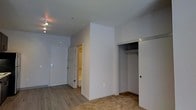 This single-floor unit offers a straightforward, functional layout designed for efficient use of space. The main living area features a combination of wood-style hard flooring near the entryway and carpet toward the interior, providing a subtle transition between zones. Clean white walls and smooth ceilings keep the space feeling bright and open.

The entryway is anchored by a solid white entry door equipped with a keypad lock, offering both security and convenience. A track lighting fixture overhead illuminates the entry zone effectively. A structural wall divider partially separates the entry from the rest of the living area, creating a natural sense of definition within the open floor plan.

Adjacent to the entry, a built-in closet with sliding doors provides practical storage, featuring a hanging rod and upper shelf — ideal for clothing, linens, or general storage needs. A secondary doorway visible beyond the divider wall leads to what appears to be an additional room or bathroom, highlighted by warm-toned walls that contrast the otherwise neutral palette of the main space.

Overall, the unit presents a clean, move-in-ready interior with a minimalist aesthetic, functional storage solutions, and a simple yet practical design that offers a solid foundation for personalization.