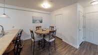 This single-floor residence offers a well-organized layout that connects living, dining, and kitchen spaces in an open, functional arrangement. The living room is anchored by hardwood-style flooring and features a sliding glass door that invites natural light and provides access to an outdoor balcony area, complemented by a side window with blinds and gray curtain panels. Reclining sofas surround a glass-topped coffee table set over a patterned area rug, creating a comfortable gathering space.

The kitchen is designed around a generous island with a built-in sink, light countertops, warm wood cabinetry, and stainless steel appliances including a gas range and refrigerator. Pendant lights hang above the island, and tile flooring distinguishes this space from the adjoining areas. A dedicated dining area nearby accommodates a round table with seating for four.

The primary bedroom features carpet flooring, a built-in wooden headboard with integrated shelving and mirror panels, and a walk-in closet. A secondary bedroom, also carpeted, offers a comfortable and understated layout. The bathroom includes warm wood vanity cabinetry, a single-sink countertop, a lighted mirror, and wood-style flooring throughout.