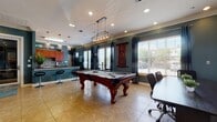 This single-floor community amenity space is thoughtfully designed to foster social interaction and relaxation across several distinct zones. The décor throughout is cohesive, defined by rich teal-painted walls, crisp white crown molding, and warm tile flooring that ties the areas together seamlessly.

At the heart of the space is a generously sized billiards area, anchored by a handsome dark wood pool table and illuminated by a statement brass-and-glass pendant light fixture. Adjacent to it, a conference-style table with rolling chairs offers a functional workspace or overflow gathering area, with large windows dressed in white blinds and teal drapes flooding the room with natural light and outdoor views.

The dining area features multiple café-style tables and chairs, bathed in natural light from floor-to-ceiling windows overlooking what appears to be a pool or outdoor courtyard. A wall-mounted flat-screen television and a cozy lounge seating area — furnished with plush tan leather sofas, an armchair, a wooden coffee table, and a decorative area rug — provide an inviting retreat for unwinding.

The kitchen is well-appointed with rich cherry wood cabinetry, granite countertops, a mosaic tile backsplash, black appliances, and a breakfast bar with barstool seating. French glass doors connect the lounge to additional areas, enhancing the open and airy flow of this versatile community space.