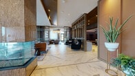 Located at 1 Fineran Way in Short Hills, New Jersey, this sophisticated lobby and amenity level showcases a refined contemporary design that blends luxurious materials with thoughtful functionality. The expansive main floor is anchored by striking chevron-patterned white marble flooring with subtle grey veining, which sets an elegant tone throughout the entire space.

The central lounge area is a standout feature, centered around a dramatic floor-to-ceiling stone fireplace surround with an integrated display screen. Curated seating arrangements — including plush sofas, accent armchairs, and low coffee tables — are thoughtfully organized on richly patterned area rugs, creating distinct yet open zones for relaxation and socializing. Warm wood ceiling panels and recessed lighting contribute to the inviting atmosphere.

Elegant bronze-framed glass partitions divide the space into multiple areas, including a more intimate lounge with deep-toned upholstered furnishings and pendant lighting. Natural light filters through floor-to-ceiling windows dressed with sheer drapery, offering views of the surrounding greenery.

Private meeting rooms, including a space labeled "Endeavor," feature bold geometric wallcoverings, patterned carpeting, and vibrant accent seating, providing functional yet design-forward settings. Lush indoor plants and curated décor accents complete this polished, hotel-caliber environment.