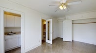 This single-floor space offers a clean and functional layout with classic design details throughout. The main room is finished with polished terrazzo flooring, which adds a timeless, durable character to the space. Crown molding runs along the upper walls, lending a subtle architectural refinement to the otherwise bright, white-painted interior. A ceiling fan with a light fixture is centrally mounted, providing both ventilation and ambient lighting. Two doorways open from the main room — one leads to a full bathroom, which is warmly lit and features a toilet, vanity with storage drawers, and a mirror, while the other provides access to a closet with a traditional paneled door, offering convenient built-in storage. The layout is straightforward and practical, making efficient use of the available space. The neutral white walls and light-toned flooring create a bright, airy atmosphere that would easily accommodate a variety of furnishing styles. Overall, the space presents a well-maintained, classic interior with functional amenities and enduring material choices.