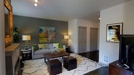 This single-floor residence offers a thoughtfully designed living space that balances modern style with comfortable functionality. The living room serves as the heart of the home, anchored by a striking dark olive accent wall that provides a rich backdrop for a vibrant, colorful piece of artwork. Dark hardwood floors run throughout, adding warmth and contrast to the light-toned walls and ceilings. The space is furnished with a plush, neutral-toned sofa accented with bold green and yellow throw pillows, centered around a rustic wooden coffee table and a textured white area rug that adds depth and softness underfoot. Leather ottoman cubes provide additional flexible seating. Ambient lighting from table lamps and ceiling-mounted track fixtures creates a warm, inviting atmosphere. Two interior doors visible from the living area lead to additional rooms, suggesting a practical and well-organized floor plan. The overall design aesthetic is contemporary and cohesive, with carefully selected décor elements that bring personality and color to the space without overwhelming it. The open sightlines and efficient layout contribute to a sense of spaciousness, making this a welcoming and visually engaging home.