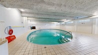 This indoor aquatic facility spans a generous single level, offering a well-equipped environment designed for both recreational swimming and relaxation. The centerpiece of the space is a sizeable rectangular lap pool with lane markers, its clear turquoise water illuminated by underwater lighting that gives the facility a bright, inviting atmosphere. A circular spa or hot tub is positioned adjacent to the main pool, accessible via stainless steel handrails and steps, making it a convenient feature for post-swim relaxation. The pool deck is finished in classic white ceramic tile, providing a clean and slip-resistant surface throughout. White-painted walls and a textured, insulated ceiling contribute to the facility's functional, enclosed aesthetic, while a row of horizontal transom windows along the upper walls allows for natural light to filter into the space. A dedicated seating area lined with white chairs offers poolside accommodation for guests or spectators, complemented by posted pool rules and safety signage. Additional amenities include a life ring mounted on the wall, ventilation ductwork overhead, and storage equipment, reflecting a fully operational and safety-conscious aquatic environment.