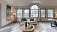 Located at 7 Southpointe Drive in Greenville, South Carolina, this single-floor space combines contemporary design with a warm, inviting atmosphere. The open layout is anchored by soaring vaulted ceilings and an impressive arched window that floods the interior with natural light, creating an airy and expansive feel throughout.

The main gathering area features large multi-pane windows framed by elegant patterned drapery, light hardwood flooring, and a comfortable seating arrangement centered around a geometric-patterned area rug. A statement fireplace with a classic white mantel serves as a focal point, accented by a bold teal feature wall and contemporary round mirrors that add depth and visual interest.

A dedicated workspace area includes built-in desks with computer stations and leather chairs, making the space fully functional for both productivity and relaxation. A live-edge wood dining table provides a natural, organic centerpiece for the gathering area, complemented by upholstered navy chairs.

Oversized abstract artwork in deep blues and golds lines the walls, lending a sophisticated artistic character. French doors with transom windows open directly to an outdoor patio, seamlessly connecting the interior to the exterior. A ceiling fan, ample floor lamps, and carefully curated décor complete this well-appointed, versatile space.