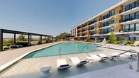 Located along River Road in Wilmington, NC, this thoughtfully designed outdoor amenity space delivers a resort-style experience with sweeping views of the surrounding waterway and natural landscape. At the heart of the space are two generously sized swimming pools with crystal-clear water, surrounded by wide pool decks appointed with lounge chairs and navy-accented umbrellas for ample sunbathing and relaxation. A covered outdoor bar area with stylish bar-top seating offers a shaded retreat directly adjacent to the pool, ideal for social gatherings. Lush artificial turf lawns provide vibrant green contrast to the sleek large-format tile hardscape throughout, while a dedicated outdoor game area features a ping-pong table set against panoramic river views. A modern pergola structure offers additional shaded seating, and an elevated terrace with a fire feature, lounge furniture, and dining tables frames unobstructed views of the waterfront. Thoughtful landscaping throughout includes ornamental grasses, native plantings, and a sculptural water feature enclosed by reed grasses and timber decking. The multi-story residential building wraps the courtyard with warm wood-tone and gray facades, private balconies, and expansive windows, creating a cohesive and vibrant community atmosphere.