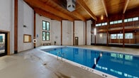 Located at 3675 Highland Ave in White Bear Lake, Minnesota, this impressive single-floor recreational facility offers a generously sized, open interior defined by striking heavy timber construction. Soaring ceilings with exposed wooden beams and tongue-and-groove wood decking give the space a warm, lodge-like character, while clerestory windows and exterior-facing windows along the walls flood the interior with abundant natural light and views of the surrounding greenery.

The centerpiece of the facility is a large indoor swimming pool, clearly marked with depth indicators and equipped with stainless steel entry rails. The pool area features a spacious surrounding deck with textured concrete flooring, ideal for safe poolside movement. Flanking the pool are rows of individual stall-style enclosures framed with metal railings, providing structured areas for attendees or program participants. An elevated mezzanine-level walkway wraps above, accessible via stairs, offering an overlooking view of the pool below.

Adjacent to the main aquatic area is a well-equipped fitness room featuring a variety of cardio and strength machines, including treadmills, ellipticals, and rowing equipment, set on wood-look flooring with recessed lighting and ceiling fans. The overall space balances functional athletic programming with architecturally rich, enduring design elements throughout.